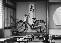 A film still of a bicycle resting against a wall and the calligraphy scroll hanging there, set apart from a larger traditional room.