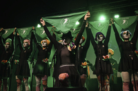Performers indoors in black costumes with colored tulle flowers and wigs hold pro-­choice green scarves above their head.