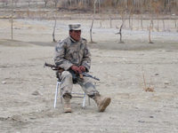 A border guard is sitting in an isolated part of the Afghan border crossing to Tajikistan in Ishkashem. It is winter and freezing. He sits on a chair with an AK-47 lying loosely across his lap. He is wearing a camouflage uniform and is clearly cold. His chair sits along an expanse of dirt, and an elderly man in a green robe is crossing the border.