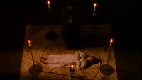 A woman lies in a pentagram, with black text chiseled inside of it.