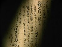 A page of parchment has black calligraphy written on it, with a sliver of light falling diagonally across it.
