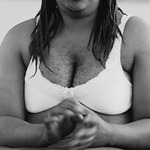Black and white portrait photograph of a black woman in white bra framed from chin to bellybutton, with prominent chest hair and hands captured in motion.