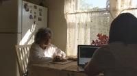 Photo: Two Black women talk while at a kitchen table in a sun-drenched room. A woman with gray hair and white blouse sits on the left. A woman with dark hair and laptop sits on the right, facing away from the viewer. They are in a kitchen with a white fridge on the left, a curtained window on the right, and a table in the near center.
