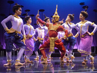 In the middle of an onstage performance, Khmer dancers in lilac dress pose in a semicircle surrounding a single masked dancer in an elaborate red and gold costume who stands with legs apart and slightly bent, the pointer finger on one hand extended to the sky.