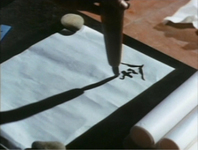 Black calligraphic characters being written on a white page, held down by stones.
