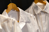 Photo of white button-down shirts hanging on wooden clothes hangers. The collars have writing on them in Arabic.