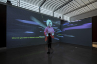 Photo: Video projected on two blue walls in a corner of a dark room. The projection is multicolored and otherworldly. Projection reads "what do you need to release to move forward?" in green. In the center of the projection is a nonbinary Black person with dark-brown skin, short white hair, and a red outfit speaking to a visitor that stands in front of the projection interacting with it.