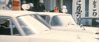 Cars await in the foreground as police officers exit a building, with calligraphy in the background.