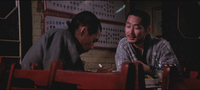 Two men converse at a table in front of banners with black and red calligraphy.