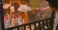 Image of a person watching a parade from a balcony. The parade contains two large lanterns with red calligraphy on them and they walk through pillars engraved in red calligraphy.