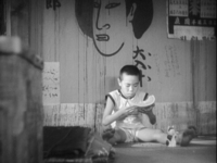 A child sits, eating melon, in front of a dirty wall covered in calligraphy, a painted face, and peeling posters.