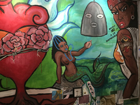 A photograph shows a mural on one of the walls of the tattoo parlor La Marca. Citlali, the Chicana superhero created by Debora Kuetzpal, appears with a tattoo of La Marca’s logo on her arm. With her defiant furled brows and her red-­colored lips half opened, Citlali’s body utters, “Say no to homophobia.” To the left of Citlali there are two images from the traditional Mexican game La Lotería: el corazón, the heart, and la sirena, the mermaid. The arrow-­pierced heart is crowned by a barbed wire with three pink carnations. With its roots digging deep in the earth, the heart emits fire from the central ventricle while wings of water spread on either side. The black Sirena holds a sea conch with her left hand. Her tight curly hair is adorned with blue beads, and she wears blue eyeshadow and blue lipstick. Photo by Mariela Méndez