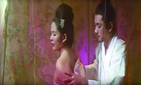 A color film still in a medium close-up shot. It shows two figures sitting in bed, covered by a transparent pinkish bed curtain. The man is holding on to the woman’s bare left arm with his left hand.