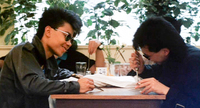 Two young grifters sit facing each other. Both wear black jackets and sunglasses. They both hold switchblades, with one holding it like a pencil carving into a wooden desk. The other is holding his switchblade forcefully by the grip, pushing it downward into the desk with his body.