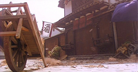 Worm's-eye view of a street with debris all around and a wagon tipped on its handle. From the roof of a building, a red and white flag waves a single large black calligraphic character.