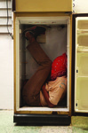 Black performer inside refrigerator, face covered by red bandana, in play about racism, poverty, disruption of community and family life.
