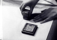 A block is pressed down by a pair of hands onto a white piece of paper, in black and white cinematopgraphy.