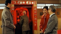 Three individuals converse while a fourth has their back to the viewer, facing a small room framed by red and gold calligraphic banners.