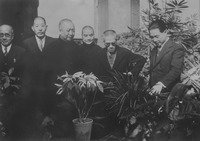 This black-­and-­white photo captures a casual moment during the conference, when the negotiators are caught on camera apparently appreciating the lush plants. Wang Jingwei, in a dark three-­piece suit and standing on the far right surrounded by plants, is pointing at an orchid while speaking. Wang Kemin, wearing tinted eyeglasses and a dark suit, and Liang Hongzhi, with shaved head and wearing a long dark robe, are both looking to where Wang is pointing. All seem rather engrossed by the conversation. Three more men in robes or suits are standing behind them. Two are looking at the plant too, while a third man on the far left is looking in the direction of the camera.