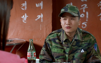 A soldier has black calligraphy printed on their jacket name badge and sits in front of white calligraphy written on the wall.