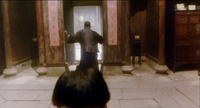 A person looks out of a doorway, away from a figure in the foreground, framed by red calligraphy scrolls.