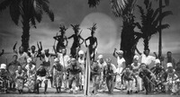 Stage scene from War Drums on the Equator showing a cast of nearly thirty Chinese actors, playing various Congolese roles, approaching the audience together. All actors are wearing black makeup applied to all portions of exposed skin. Actors playing Congolese guerilla fighters are toting machine guns and spears, and appear with bare chests and feet. Palm trees form the background scenery.
