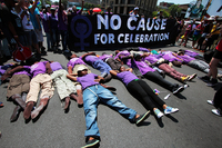 Six black women sprawl across asphalt, surrounded by straw-stuffed dummies, backed by a banner reading “No Cause for Celebration,” alongside a purple “women’s symbol” with fist embedded in its center.