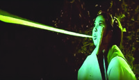 A color still from a film. It shows a female figure emitting out a laser-like greenish light from her month.