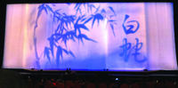 A color photo of a stage curtain in a theater. It shows a milky white silk certain with two characters in Chinese calligraphy to the right and to the left, Chinese ink paintings of bamboo leaves and branches.