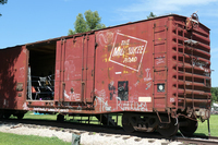 Fig. 32 A rust-colored boxcar, covered with graffiti, stands on railroad tracks in a grass field representing a hobo jungle. The boxcar door is open, revealing lines strung for hanging laundry, along with an orange chair inside.