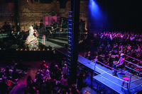 A wrestling ring set up and surrounded by audience members in the cavernous space of St. Ann’s Warehouse during a performance of A 24-­Decade History of Popular Music in Brooklyn, New York.