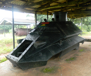 The black metal shell of an armored vehicle stands on a concrete floor under a corrugated iron roof. The room in which it is housed has no walls, and beyond can be seen grass-­covered ground, several trees, and another museum shelter that contains a metallic storage tank. The Biafran Red Devil armored vehicle is angular and warlike, with a large rotating turret at the top. A Biafran flag is painted at the top of the vehicle’s front face.