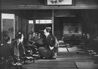 A film still of a man kneeling in the center of a room with an arm out to a row of men at left, with his back to a mostly off-screen row of men at right. A screen with calligraphic text appears behind the left row of men and far in the back of the room.