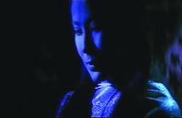 A color film still in a medium close-up shot. It shows a woman’s face illuminated by a dark blue light. She is wearing a discreet smile on her face, and her robe appears to be translucent.