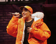Solo performer wearing a black t-­shirt with an orange parka and a baseball cap atop a gray do-­rag. She holds a microphone and addresses the audience with her hand touching her chest.