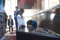 Mourners pay homage at Hojaji’s cenotaph in his home town, Najaf Abad (Isfahan Province, Iran). A man in a blue suit kneels, head bowed into his hands, which rest on the cenotaph’s top. The cenotaph is large, flat, and slightly inclined, black marble with white writing in Farsi. A boy stands beside him, and several mourners stand behind. The cenotaph is located in a tall room with patterned rugs across the floor and simple, geometric decoration across the walls.