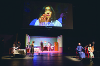 Figure 5.1: A color photograph of a theater stage with camera operators, actors, and musicians and an overhead projection screen showing a close-­up of a woman’s face.