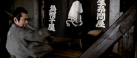 Yojimbo is bares his sword in front of a hat shop with a calligraphic noren.