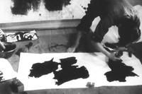 A photo of a calligrapher leaning over a large white piece of paper, drawing thick calligraphic text. At the left of the photo, are additional brushes, bowl of ink, and other supplies.