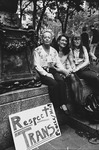 Three transgender women sitting outdoors in an urban park. Hand-drawn poster says “Respect Trans People/Men!”