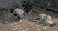 Three men squat over calligraphic characters carved into a wood floor, painting them in various colors. A fourth man sleeps on the floor in the lower right.