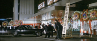 Calligraphy is visible on signs and banners as men bow to a black car arriving at the building.