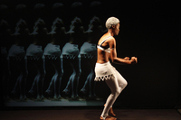 Fig. 10. Solo black female dancer in front of screen mirroring her image. Dancer in white lace skirt, white tights, bra, and powdered hair. Moving stage left, arms bent at elbows.