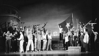 Stage scene in which a cast of roughly twenty Chinese actors playing various Vietnamese roles celebrate victory by raising their arms, guns, and a North Vietnamese flag. To the left, a Vietnamese character points a machine gun at a surrendering U.S. officer. A ship labeled “U.S. Army” forms the background, next to a rising sun.