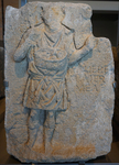 A weathered stone relief depicting a man standing, facing forward. He wears a tunic, tied at the waist, and carries a basket that hangs from his neck; within the basket are rounded shapes. The figure’s hands are raised, but too damaged to be interpreted.