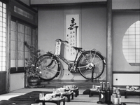 A tokonoma with a striking "kotobuki" on a scroll; the vertical stroke is just off-center. It hangs behind a bike, which has a tag hanging from the handlebars with calligraphy. In the foreground are vacant mats and short tables with dishes and bottles arranged.