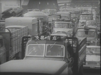 A truck in traffic has a sign at the top of its windshield with white calligraphy printed on it, in black and white cinematography.