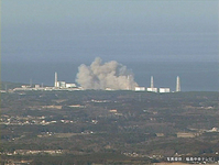 A color video still, showing a cloud of grey smoke rising against a grey-blue sky from a series of white buildings and towers in the far distance. A few smaller buildings and many trees can be seen throughout the hills in the foreground.