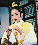 A color film still in a medium shot. It features a female figure with her long black hair pulled up in a bun with beautiful yellow decorations. She wears a white robe with matching yellow decorations along her long collars and long sleeves. She is raising a small cup with both hands.
