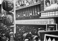 A black and white photo of an old theater, with a crowd gathered in front. Calligraphic text covers the numerous boards over the entrance way and the paper lanterns in front.